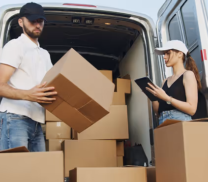 Man wearing a black cap carrying a cardboard box and woman in a white cap checking a tablet near an open delivery van filled with boxes.
