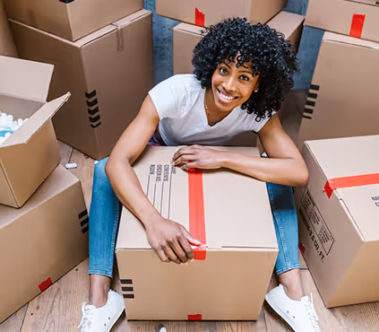 Smiling woman sitting on the floor holding a cardboard box, surrounded by other packed boxes.
