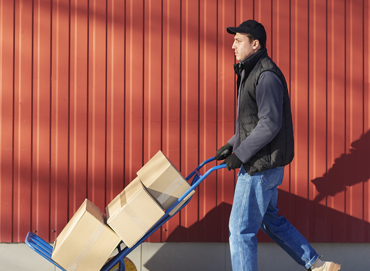 Delivery worker in a black cap, vest, and gloves pushing a blue hand truck carrying three cardboard boxes along a red metal wall.