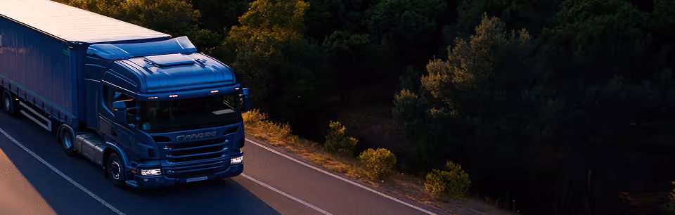 Blue semi-truck driving on a road alongside a forested area during sunset.