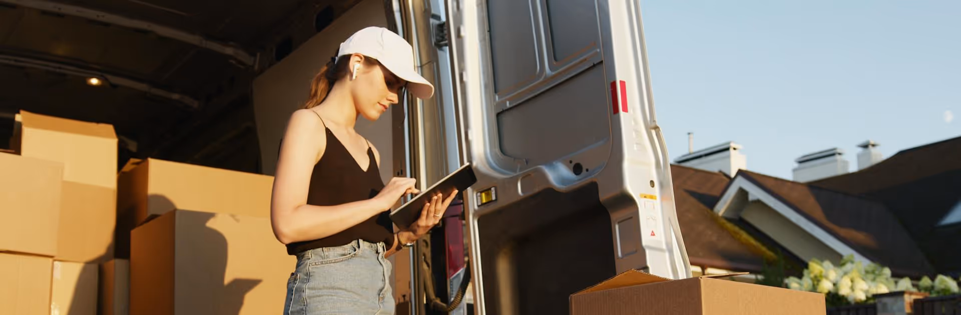 Young woman wearing a white cap and earbuds, using a tablet in front of a van loaded with cardboard boxes.