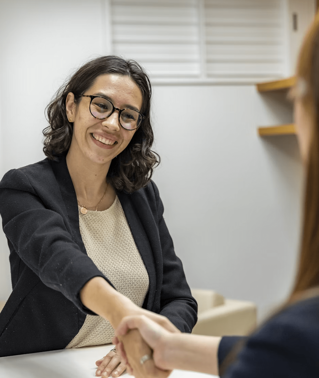 Smiling woman wearing glasses and business attire shaking hands with another person in an office setting.