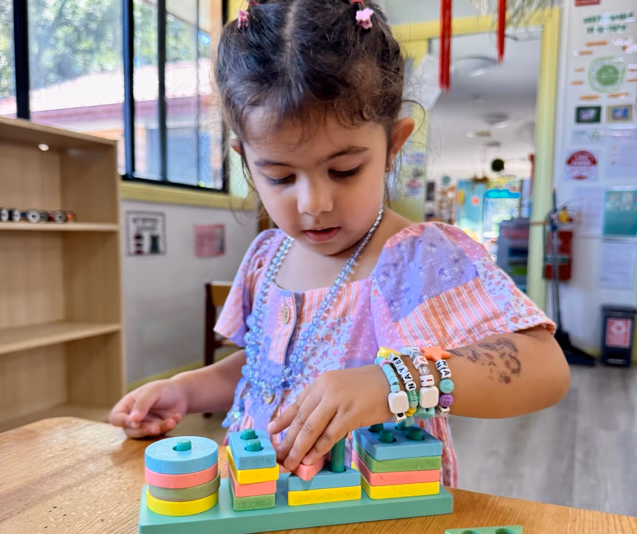 Young girl wearing colorful bracelets and a purple dress playing with a wooden stacking toy at a table in a classroom.