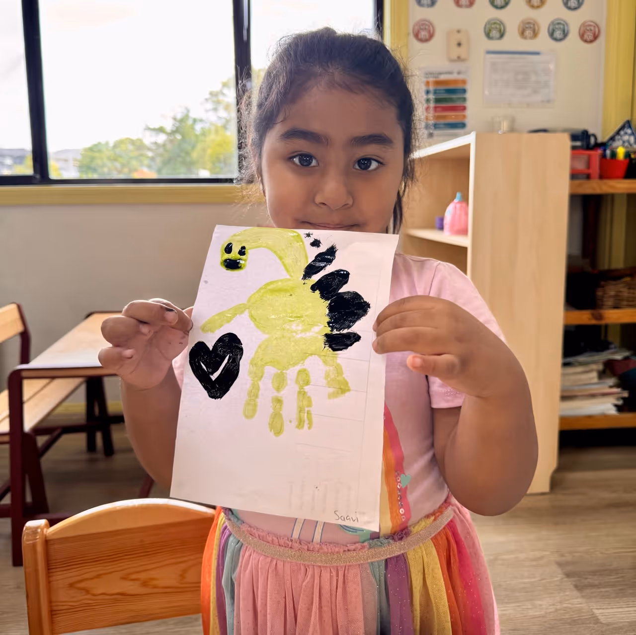 Young girl holding a drawing of a yellow and black dinosaur with a black heart.