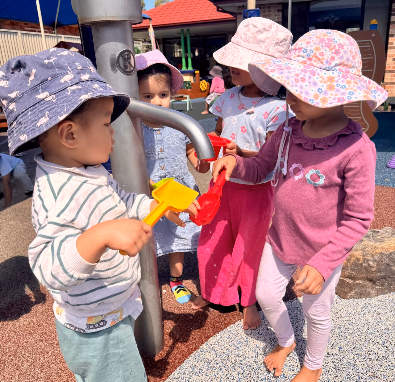 Four young children wearing sun hats playing with water and colorful shovels at an outdoor playground water pump.