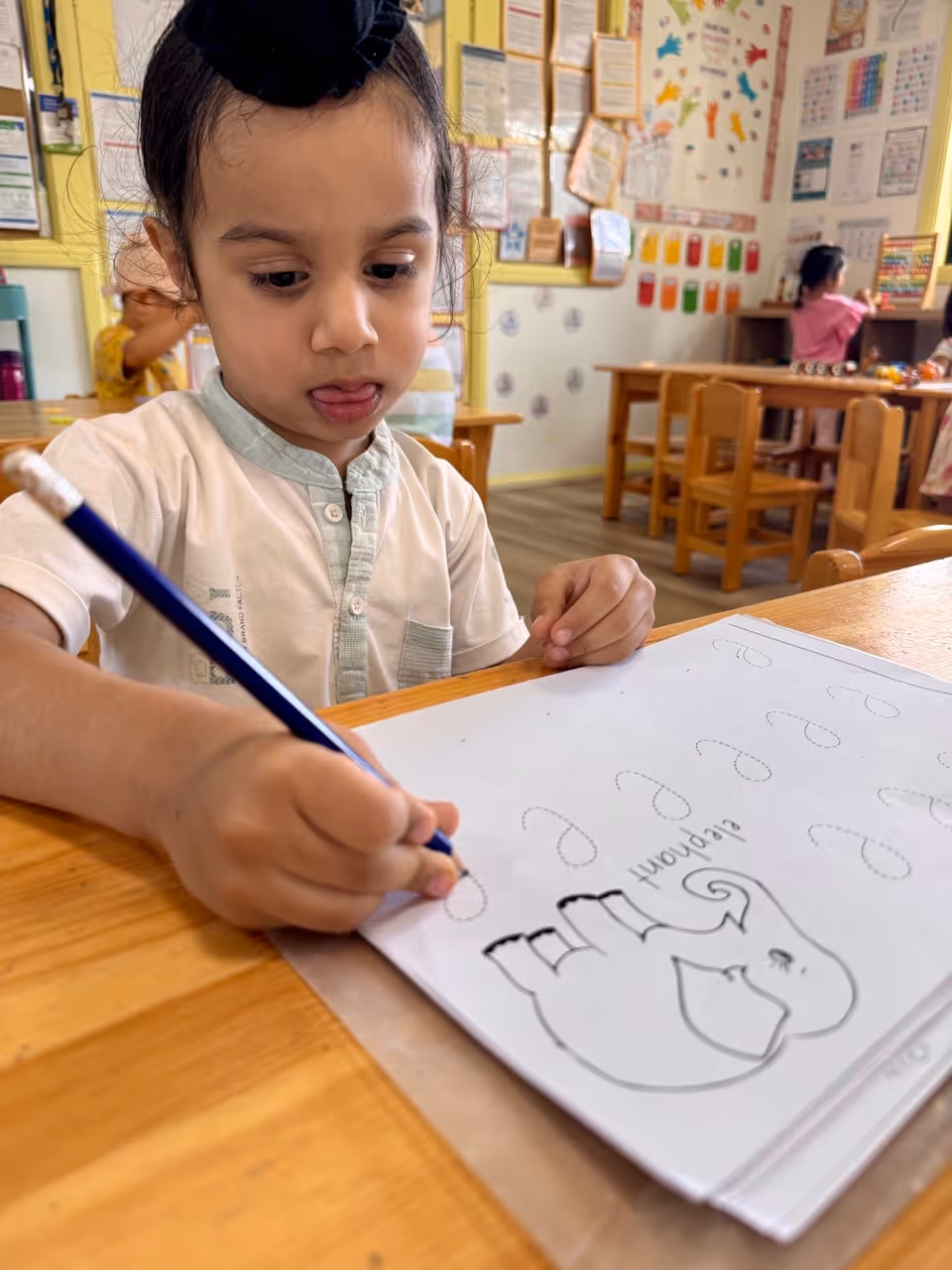 Young girl coloring a picture with colored pencils at a classroom table with a boy and educational posters in the background.