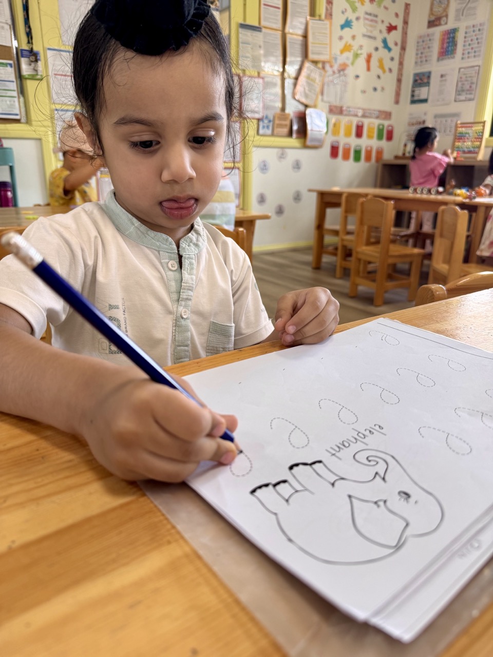 Young girl coloring a picture with colored pencils at a classroom table with a boy and educational posters in the background.