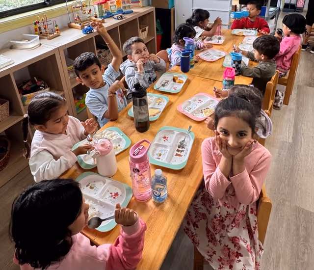 Group of young children sitting around a wooden table eating meals from divided trays in a brightly lit classroom.