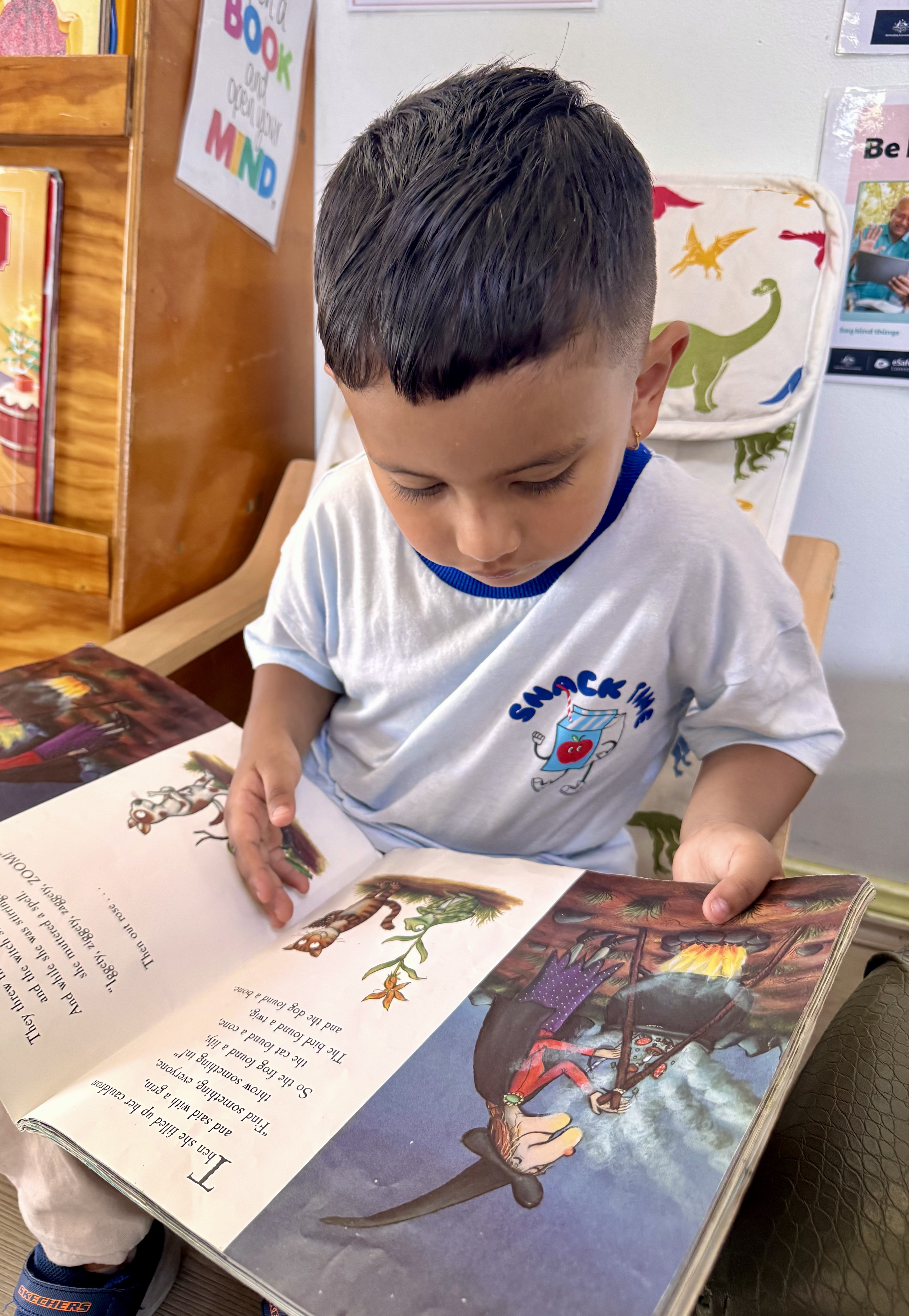 Young girl coloring a picture with colored pencils at a classroom table with a boy and educational posters in the background.