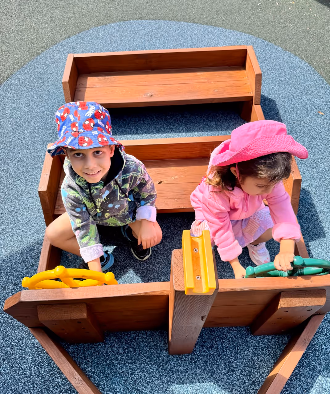Young girl coloring a picture with colored pencils at a classroom table with a boy and educational posters in the background.