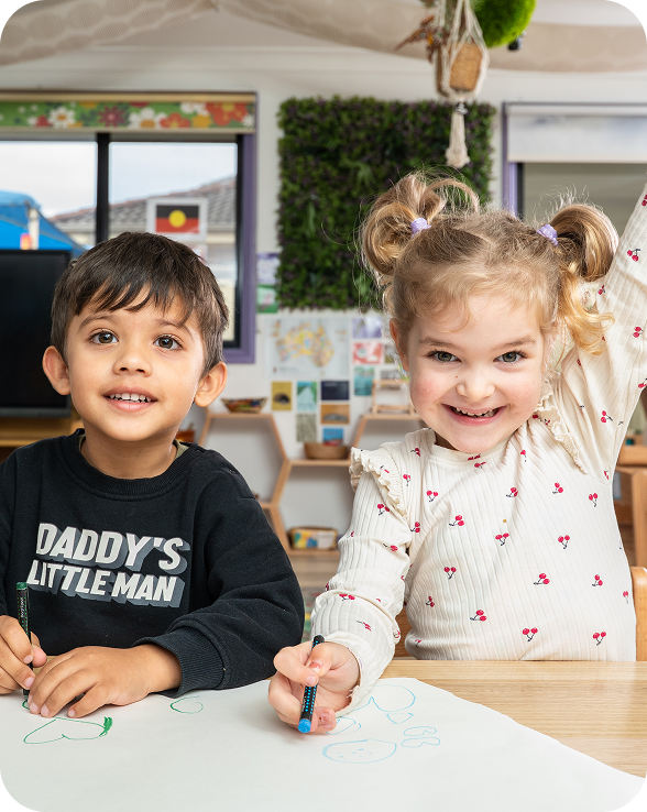 Young girl coloring a picture with colored pencils at a classroom table with a boy and educational posters in the background.