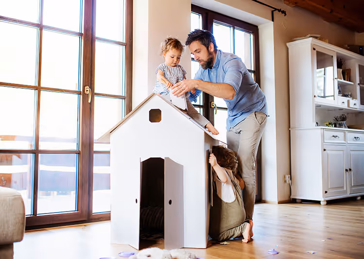 Father playing with two young children around a cardboard playhouse in a sunlit living room.