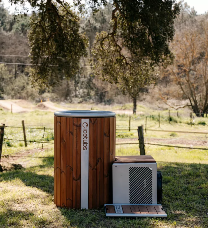 Icetubs wooden hot tub and accompanying mechanical unit on grass under a large leafy tree in a rural outdoor setting.