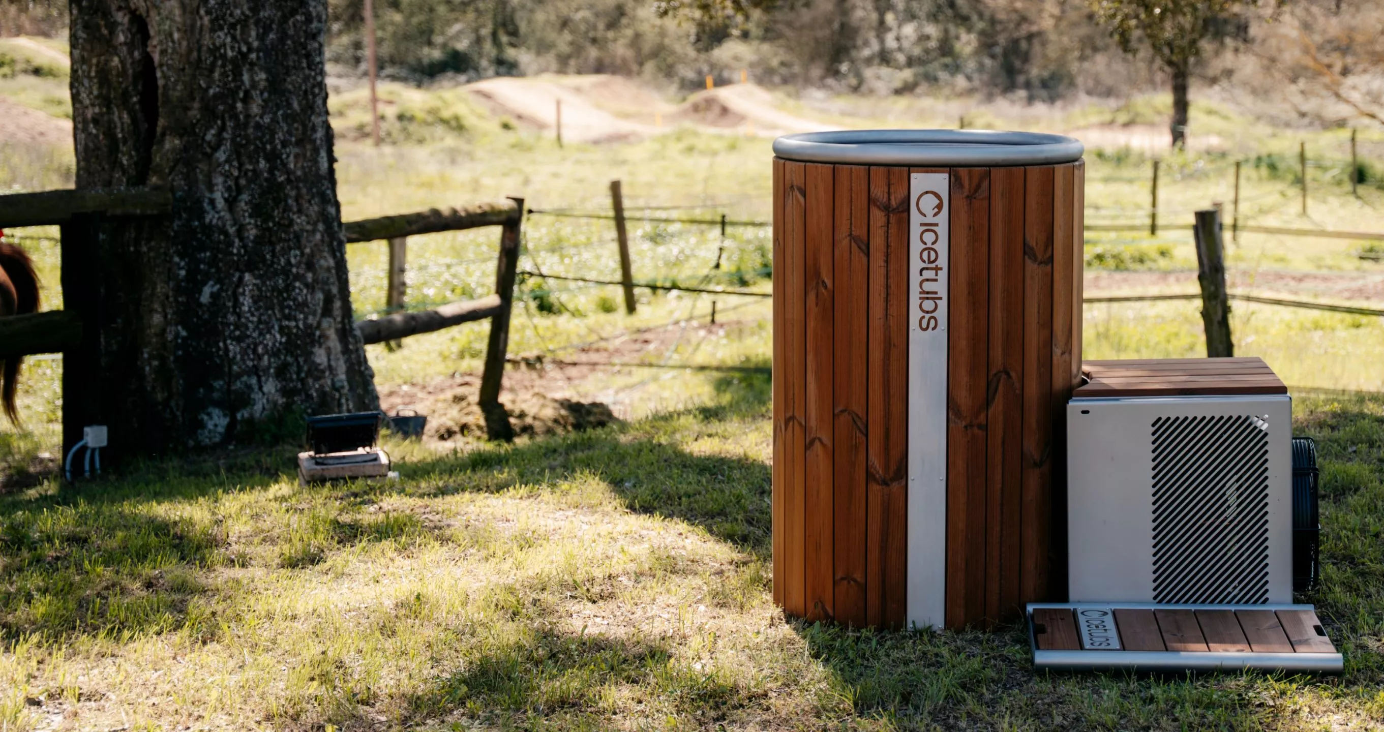 Wooden hot tub with metal accents branded 'Icetubs' placed outdoors on grass near a tree and rustic wooden fence.