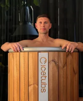 Man standing inside a wooden ice tub sauna, holding the tub's rim with both hands.