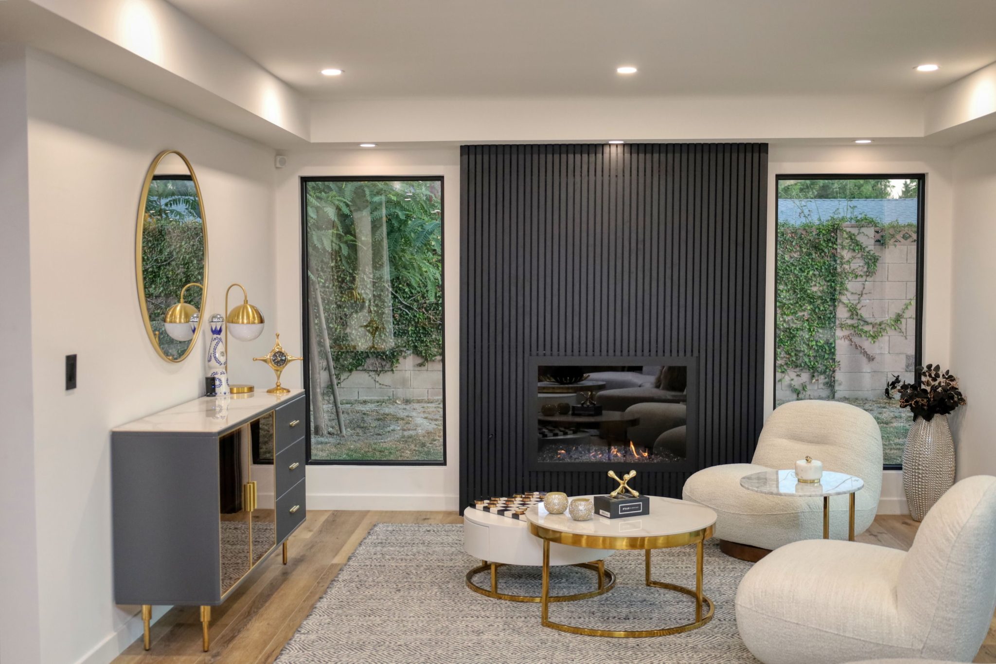 Modern living room with dark fluted fireplace wall, cream curved chairs, gold-accented coffee table, and large windows overlooking garden