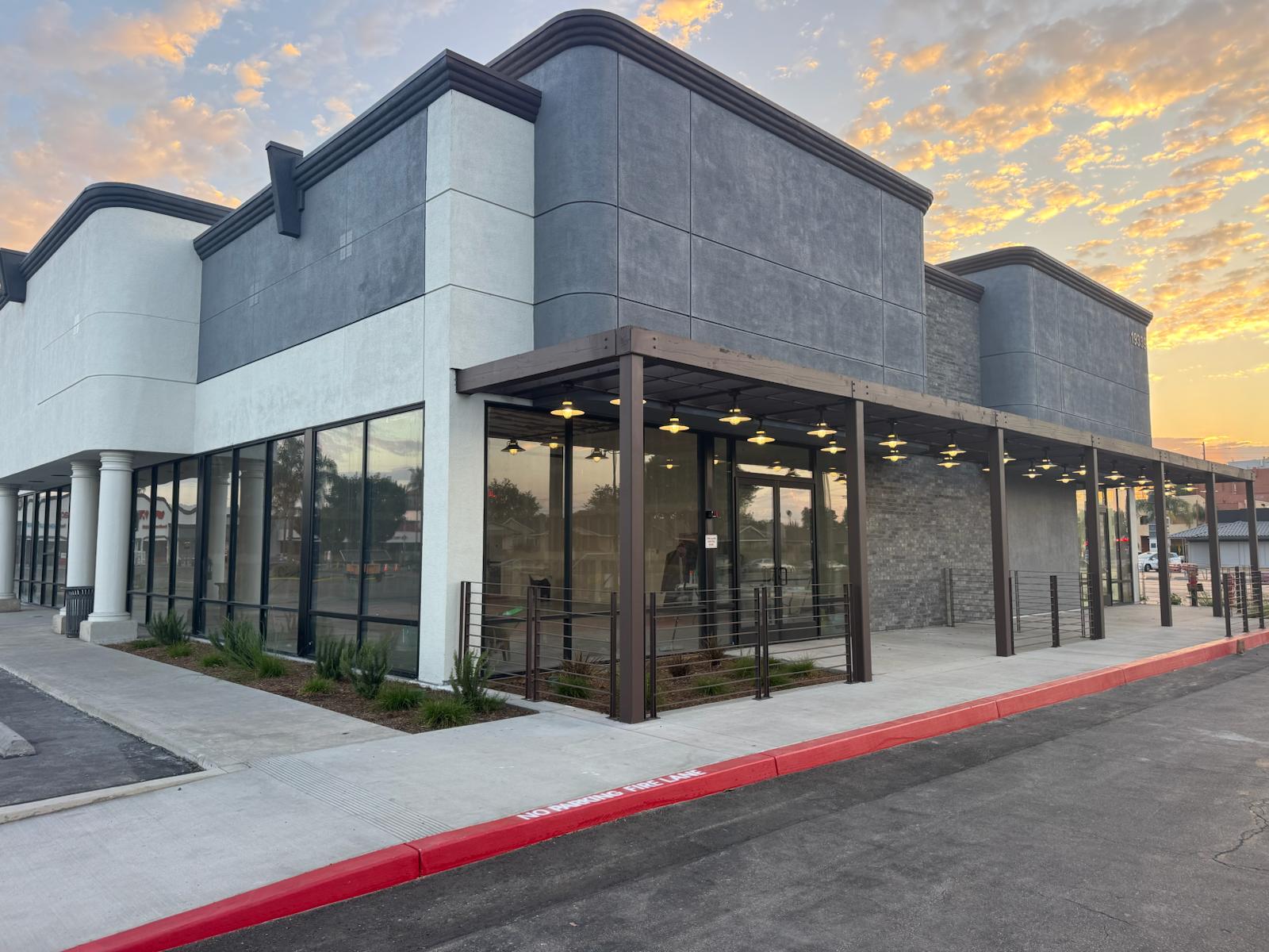 Modern commercial retail building with gray stone and white stucco exterior, floor-to-ceiling windows, and covered walkway at sunset