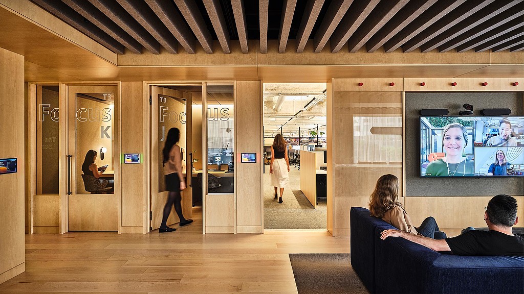 Modern office interior with light wood paneling, glass-walled Focus rooms, exposed beam ceiling, and employees in collaborative workspace