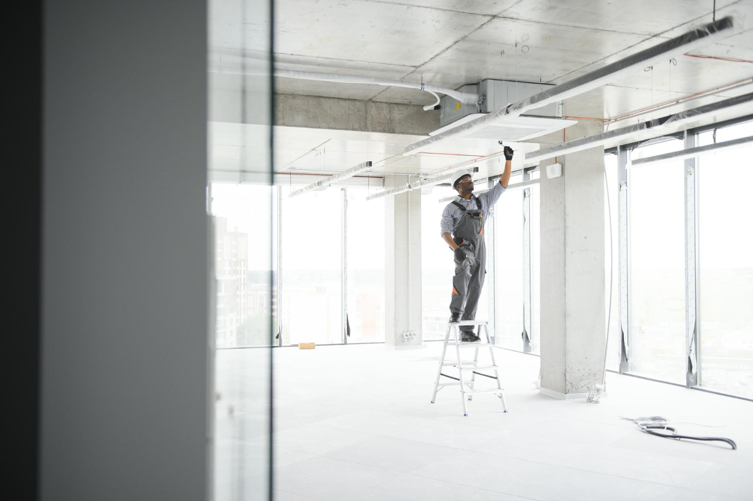 Construction worker in gray coveralls on ladder installing ceiling components in modern commercial building interior