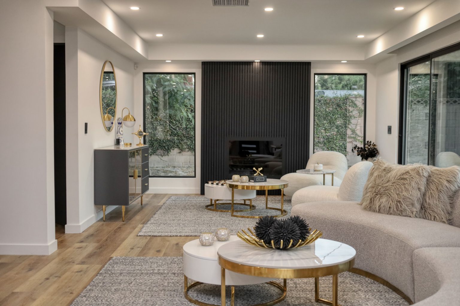 Modern living room with beige sectional sofa, gold accent tables, dark fluted fireplace wall, and large windows overlooking greenery