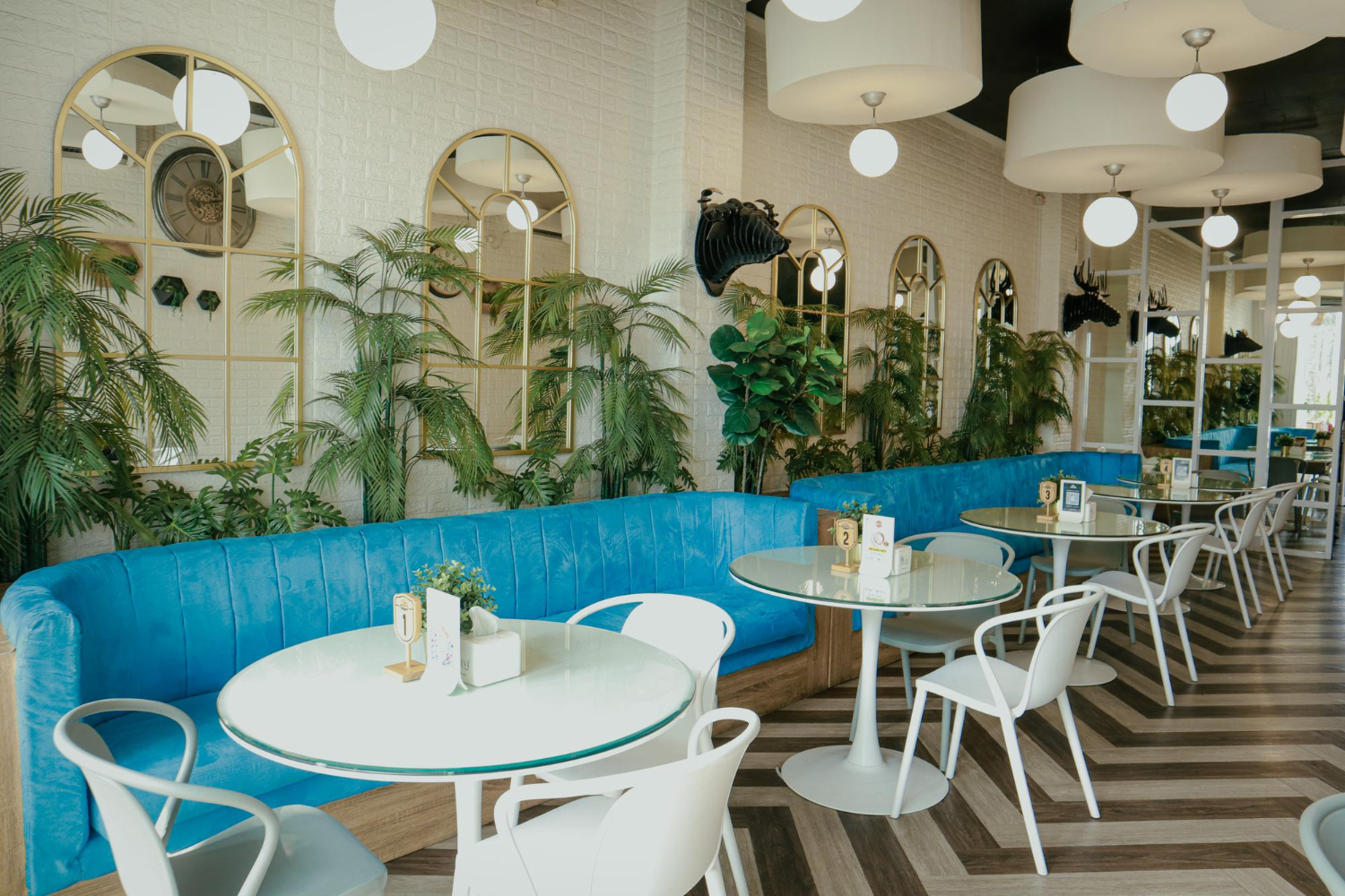 Modern restaurant interior with bright blue curved banquettes, white round tables, gold-framed arched mirrors, and lush tropical plants