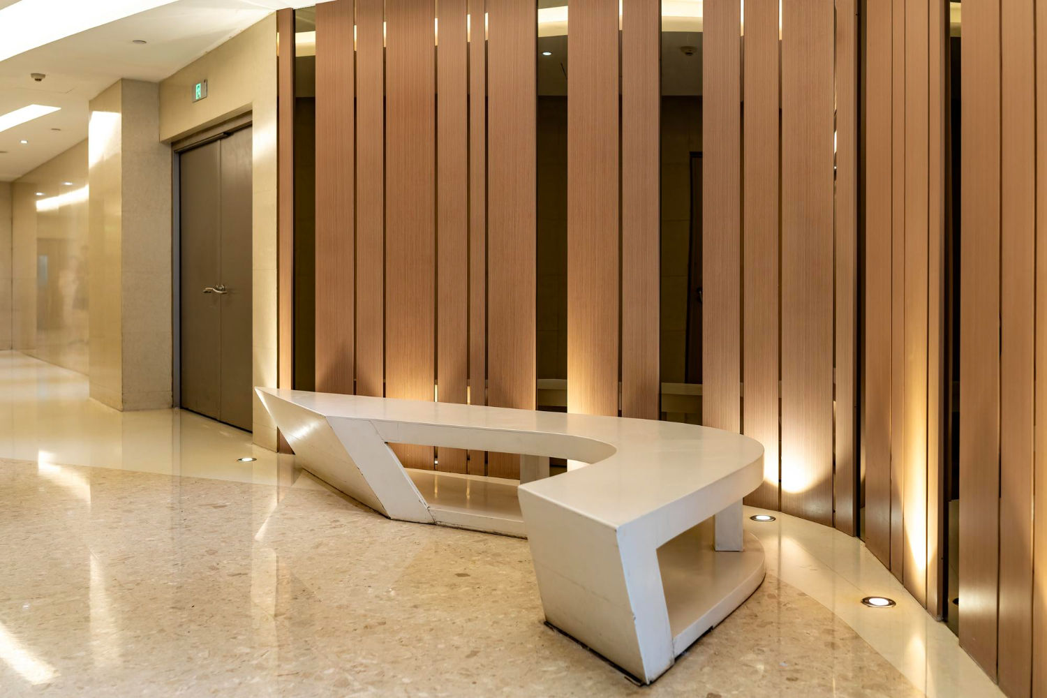Modern commercial lobby with white sculptural bench, warm wood slat wall panels, polished marble floors, and uplighting