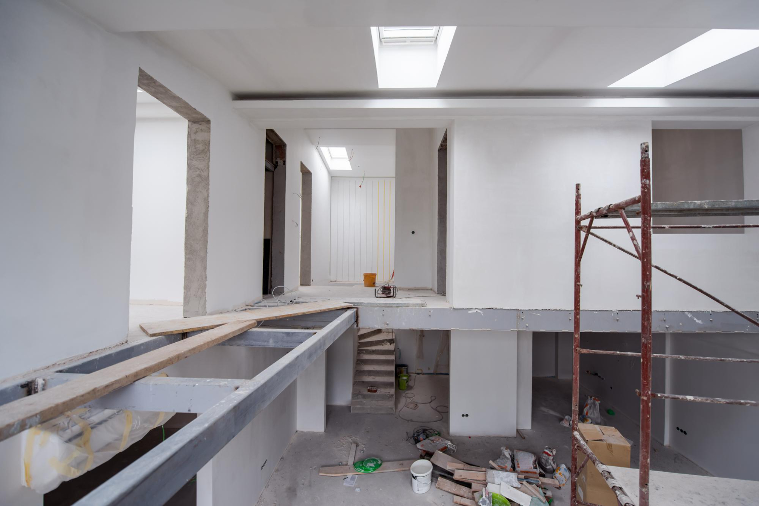 Interior office renovation in progress with white walls, exposed concrete beams, red scaffolding, and skylights overhead