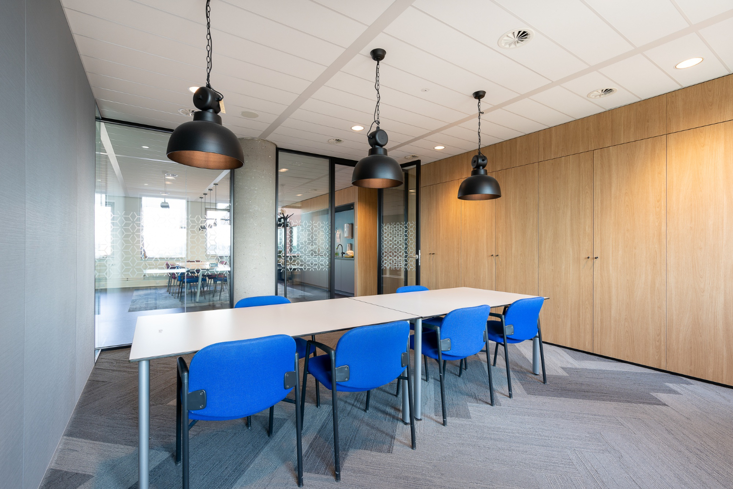 Modern conference room with white table, blue chairs, black pendant lights, wood wall panels, and glass partitions