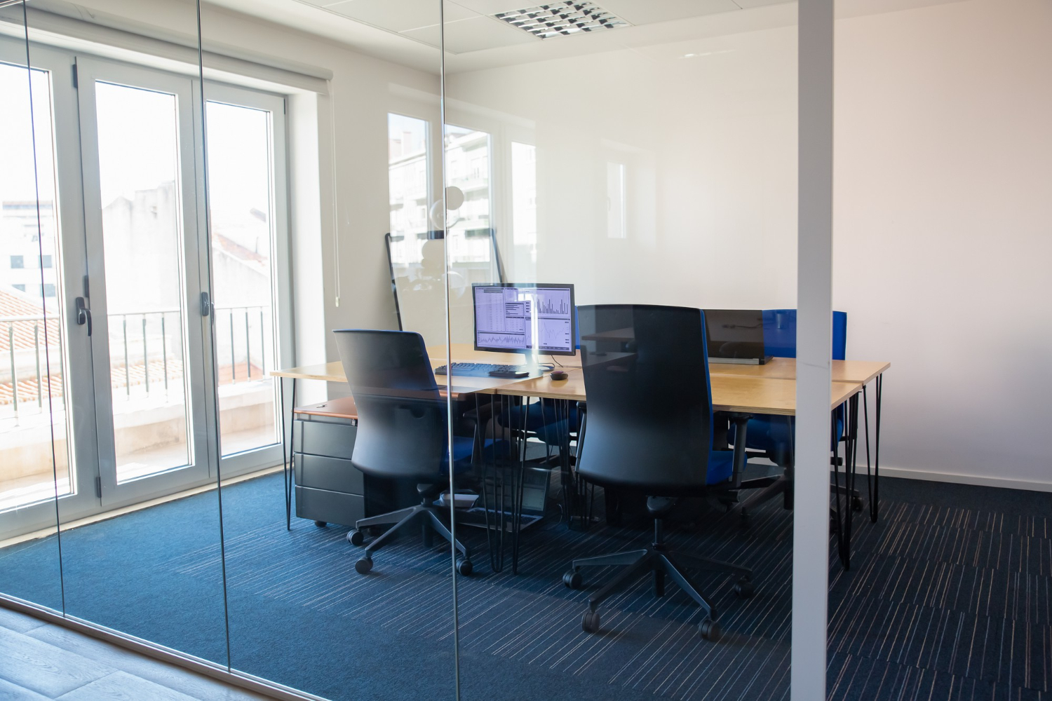 Modern glass-walled conference room with wood table, black chairs, computer monitors, and balcony doors in renovated office