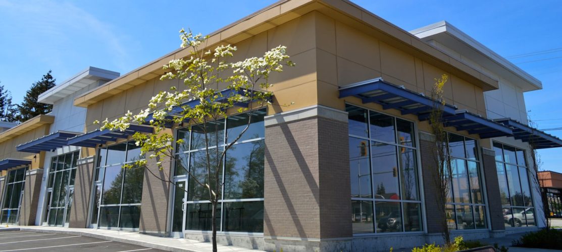 Modern commercial building with glass storefronts, beige brick exterior, metal awnings, and contemporary architectural design