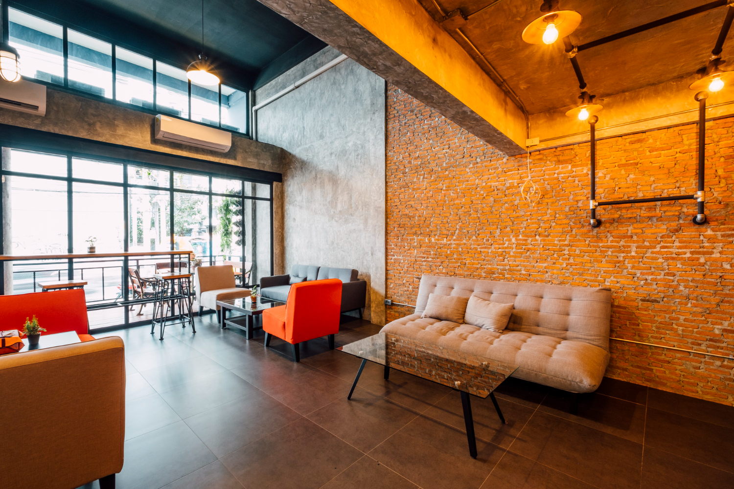 Modern commercial interior with exposed brick wall, concrete ceiling, large windows, and mixed seating with orange and beige furniture