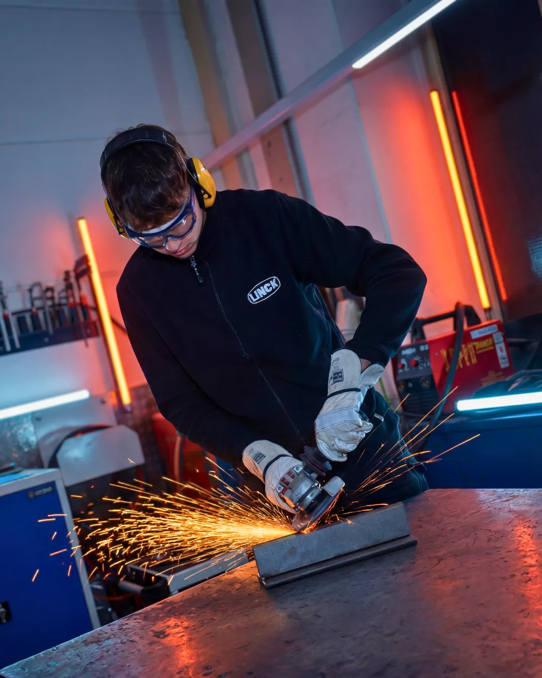 Ein Auszubildender im schwarzen LINCK-Pullover bearbeitet ein Bauteil mit einem Winkelschleifer, während Funken sprühen.