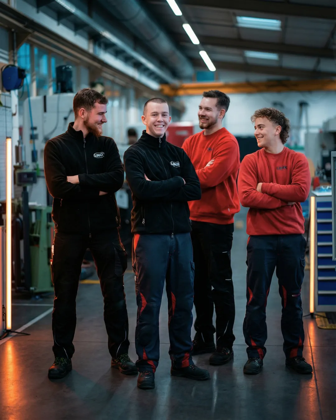 Vier männliche Auszubildende stehen nebeneinander in der Ausbildungswerkstatt und lachen in die Kamera. Im Hintergrund ist orangefarbene Neonbeleuchtung zu sehen.