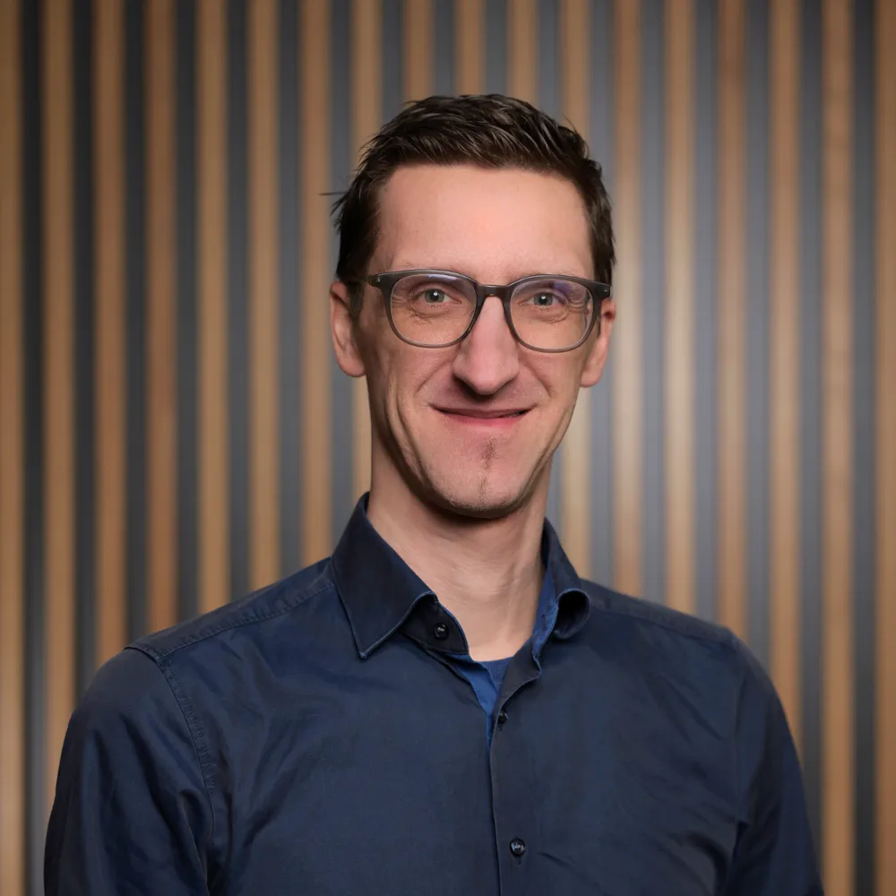Personalleiter Daniel Gaus lächelt in die Kamera, vor einem Hintergrund mit hölzernem Akustikpanel.