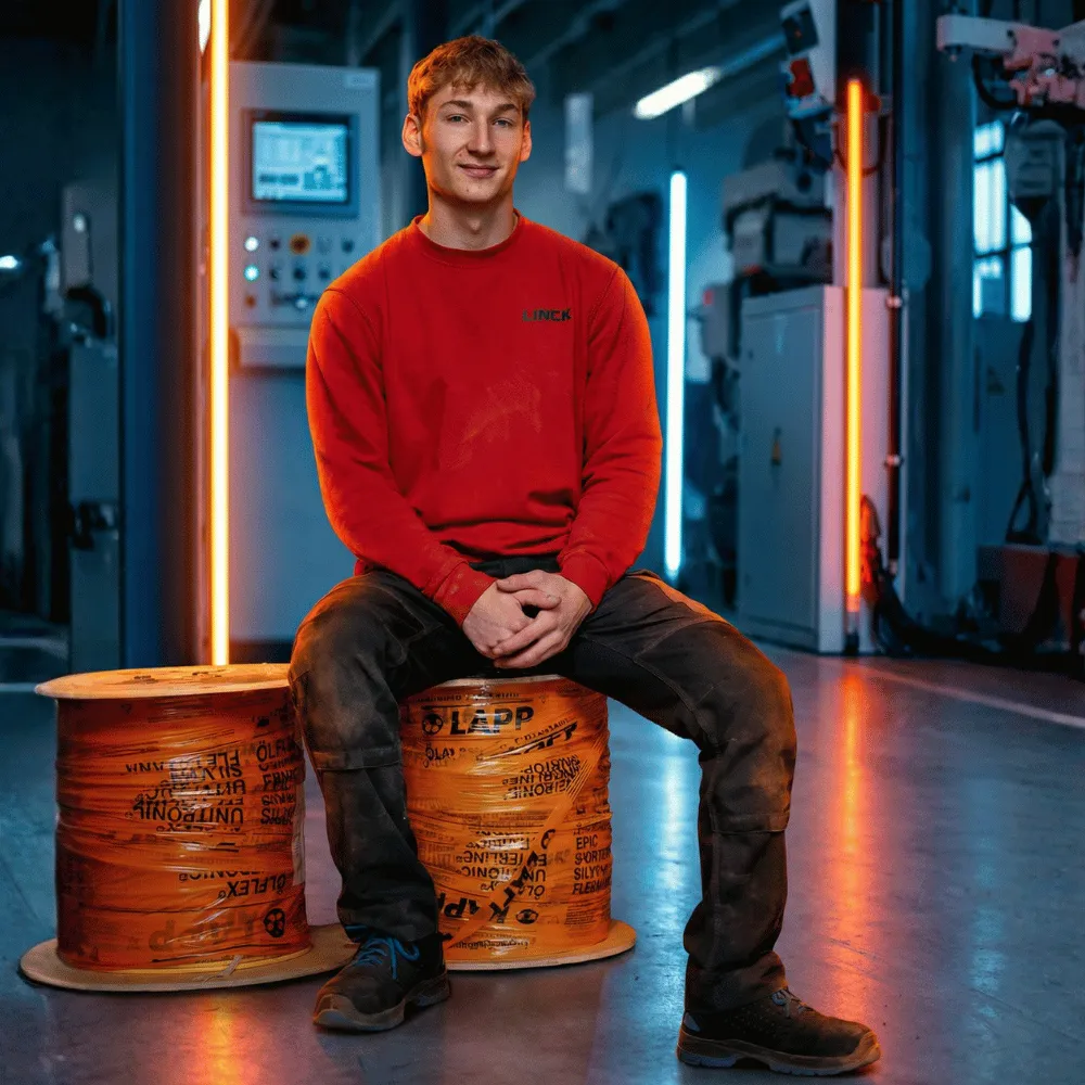 Ein Auszubildender sitzt lächelnd auf einer orangefarbenen Kabeltrommel. Im Hintergrund sind eine Maschine und blau-orange Neonbeleuchtung zu sehen.