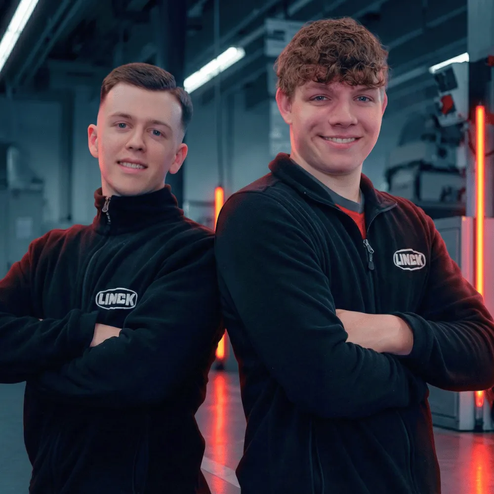 Zwei LINCK-Auszubildende stehen Rücken an Rücken mit verschränkten Armen in der Werkshalle und lachen in die Kamera