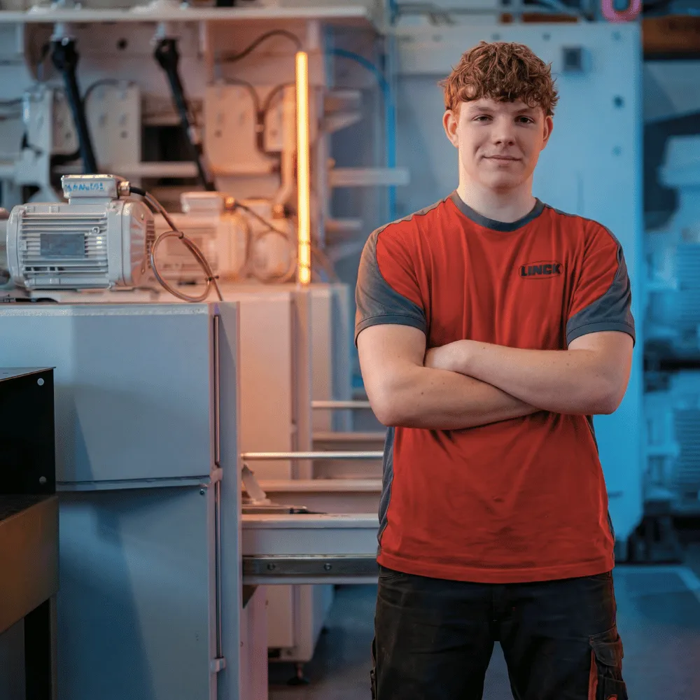 LINCK-Auszubildender im roten Shirt steht mit verschränkten Armen lachend vor einer Maschine