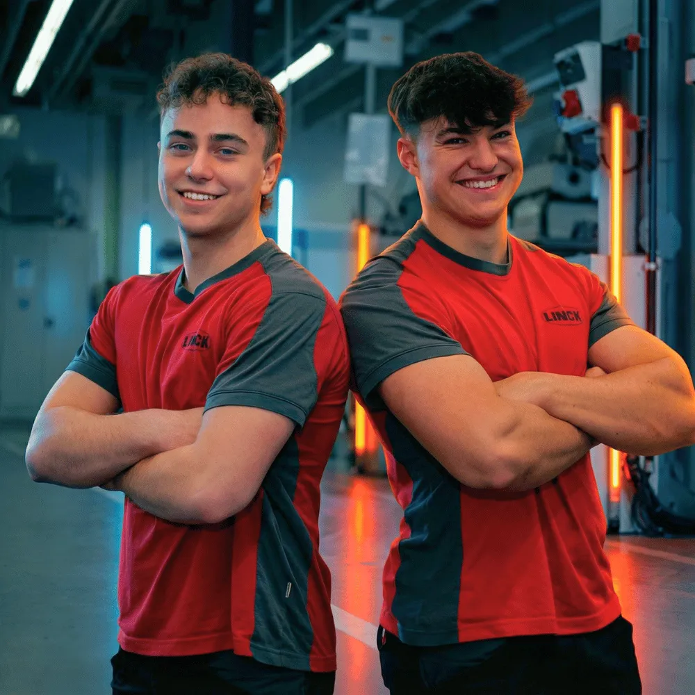 Zwei LINCK-Auszubildende stehen Rücken an Rücken mit verschränkten Armen in der Werkhalle und lächeln in die Kamera