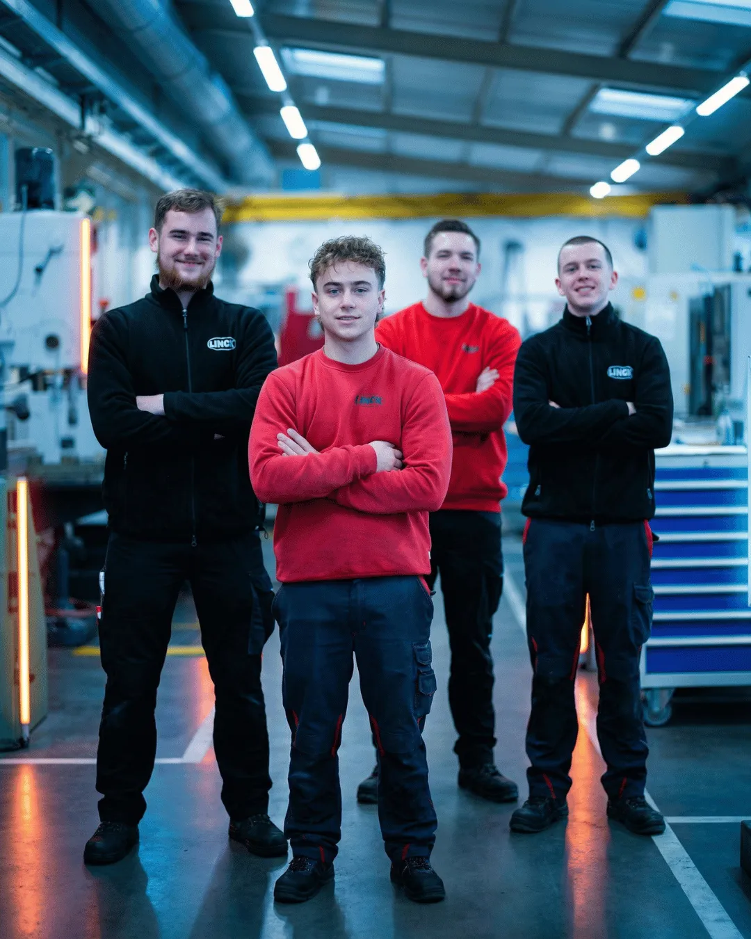 Vier LINCK-Auszubildende stehen in der Ausbildungswerkstatt versetzt nebeneinander, tragen rote und schwarze LINCK-Pullis und lachen mit verschränkten Armen in die Kamera