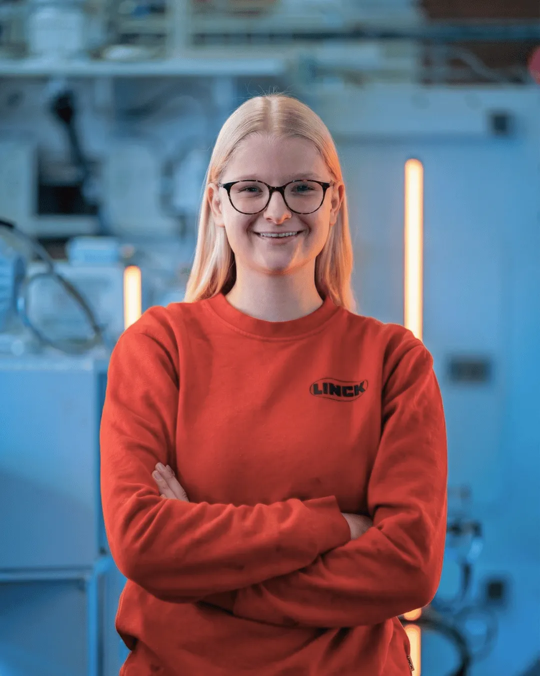 LINCK-Auszubildende im roten Pulli steht mit verschränkten Armen vor einer weißen LINCK-Maschine und lächelt in die Kamera