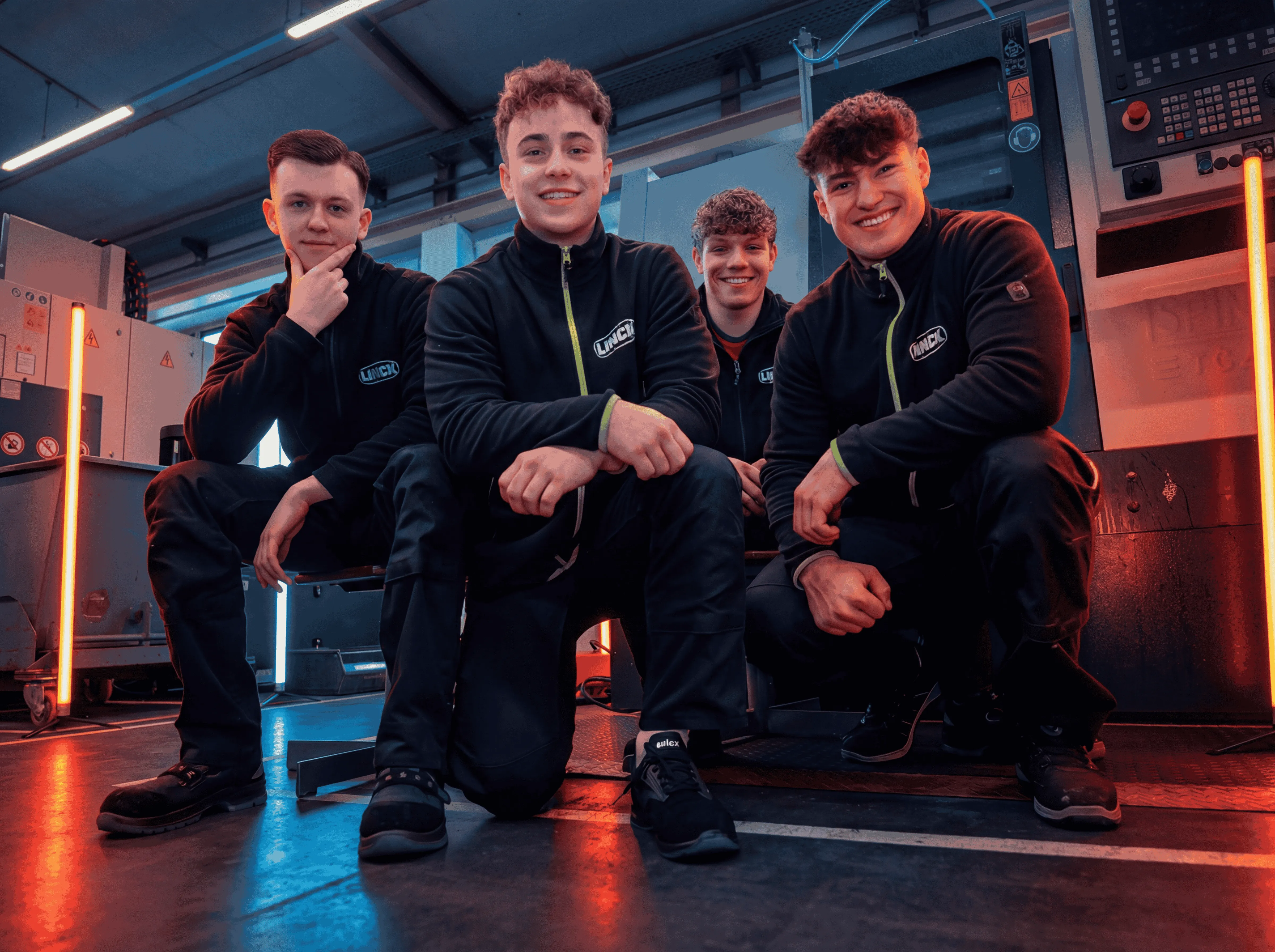 Vier LINCK-Auszubildende im schwarzen Pullover knien auf dem Boden, lachen in die Kamera und posieren vor Maschinen und orangefarbenen Neonröhren