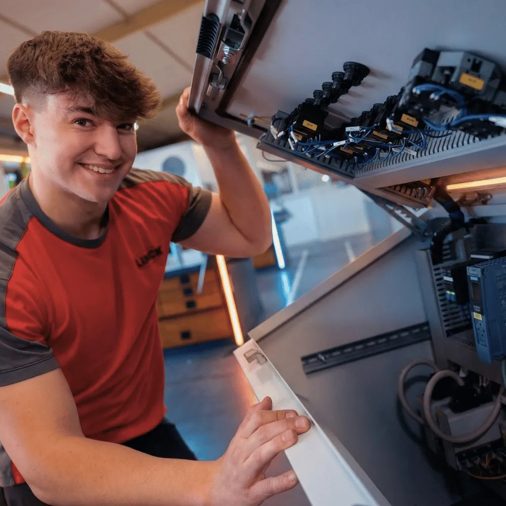 LINCK-Auszubildender im roten Shirt öffnet einen Stromkasten in der Ausbildungswerkstatt und lächelt in die Kamera