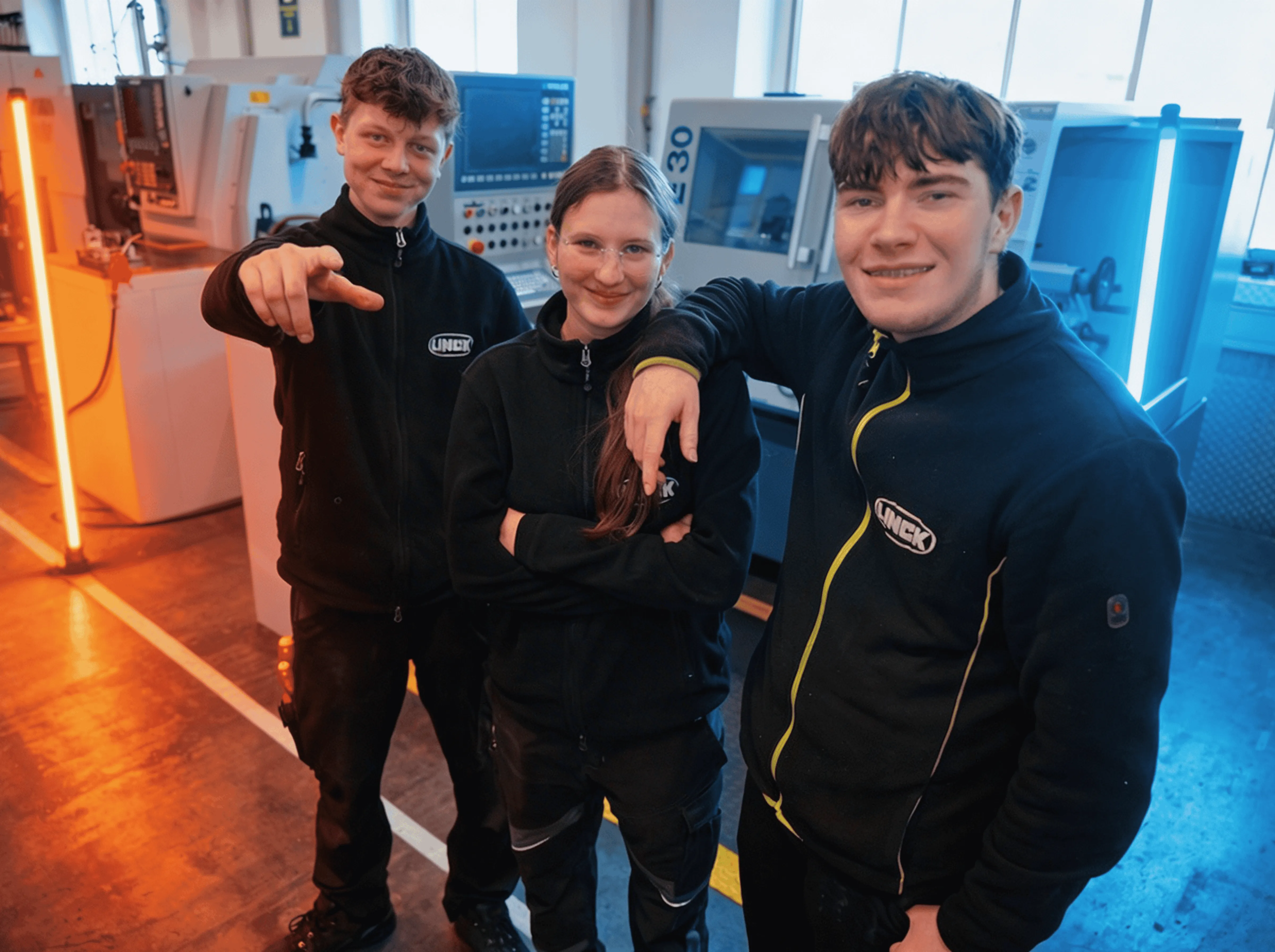 Drei LINCK-Auszubildende im schwarzen Pulli stehen in der Ausbildungswerkstatt, posieren lächelnd in die Kamera und werden von orangefarbenen und blauen Neonröhren im Hintergrund begleitet