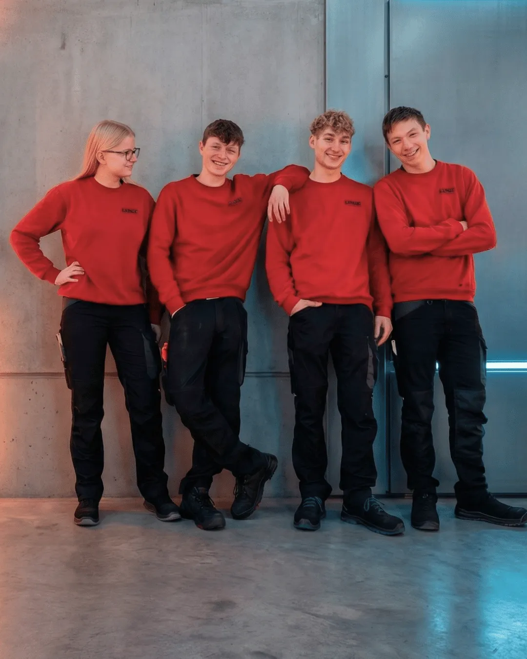 Auszubildende von LINCK posieren an einer grauen Wand in roten Pullis und schwarzen Arbeitshosen, während drei in die Kamera blicken und eine Person lachend zu den anderen schaut