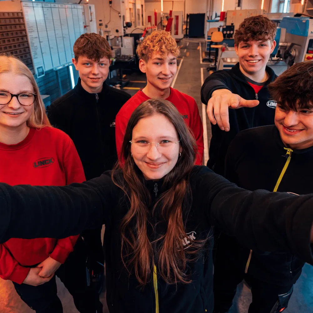 Sechs LINCK-Auszubildende blicken in die Kamera, während die Person im Vordergrund ein Selfie in der Ausbildungswerkstatt mit orangefarbenen Neonröhren aufnimmt
