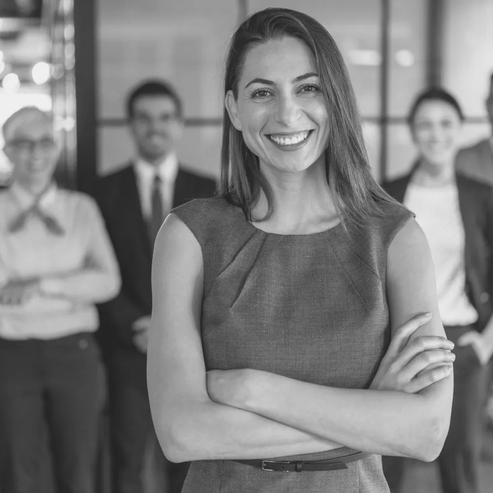 Selbstbewusste Frau im grauen Etuikleid mit verschränkten Armen blickt lächelnd in die Kamera, im Hintergrund unscharfe Silhouetten von drei Personen