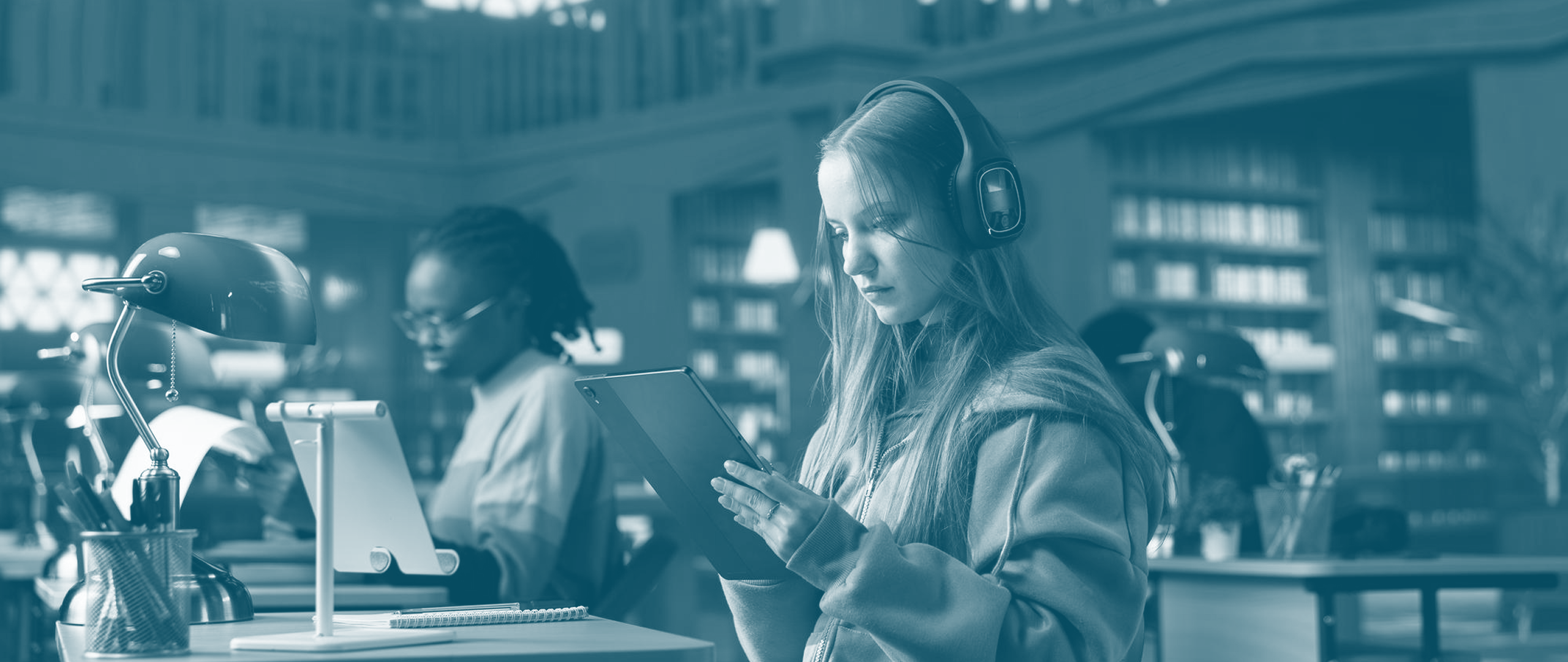 Young woman wearing headphones and reading a tablet in a library study area with another person working in the background.