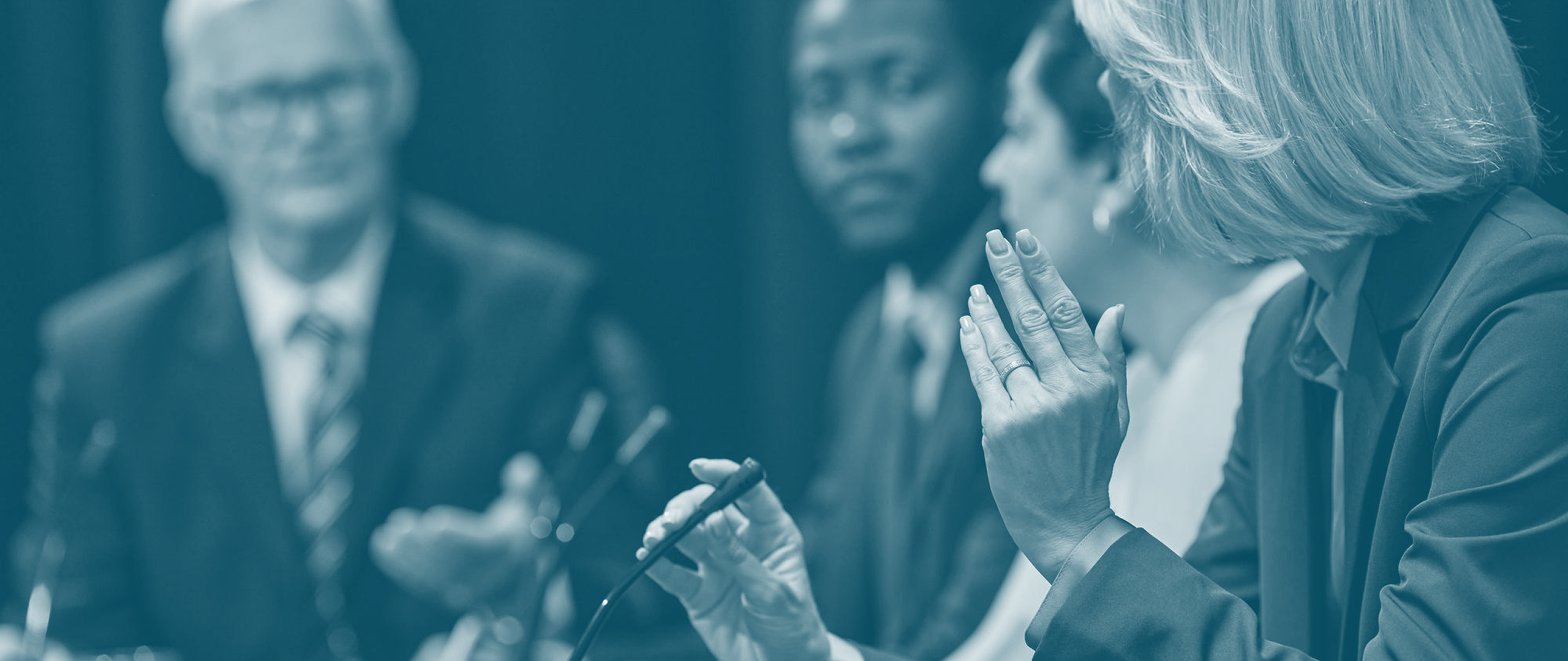 Business panel discussion with four professionals seated, one woman speaking into a microphone and gesturing with her hand.