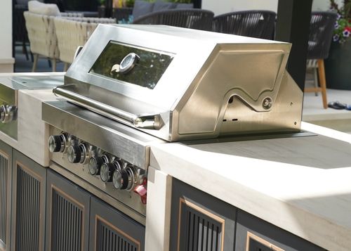 Stainless steel built-in outdoor grill with control knobs and thermometer on the lid, set in a white stone countertop with dark cabinets below.