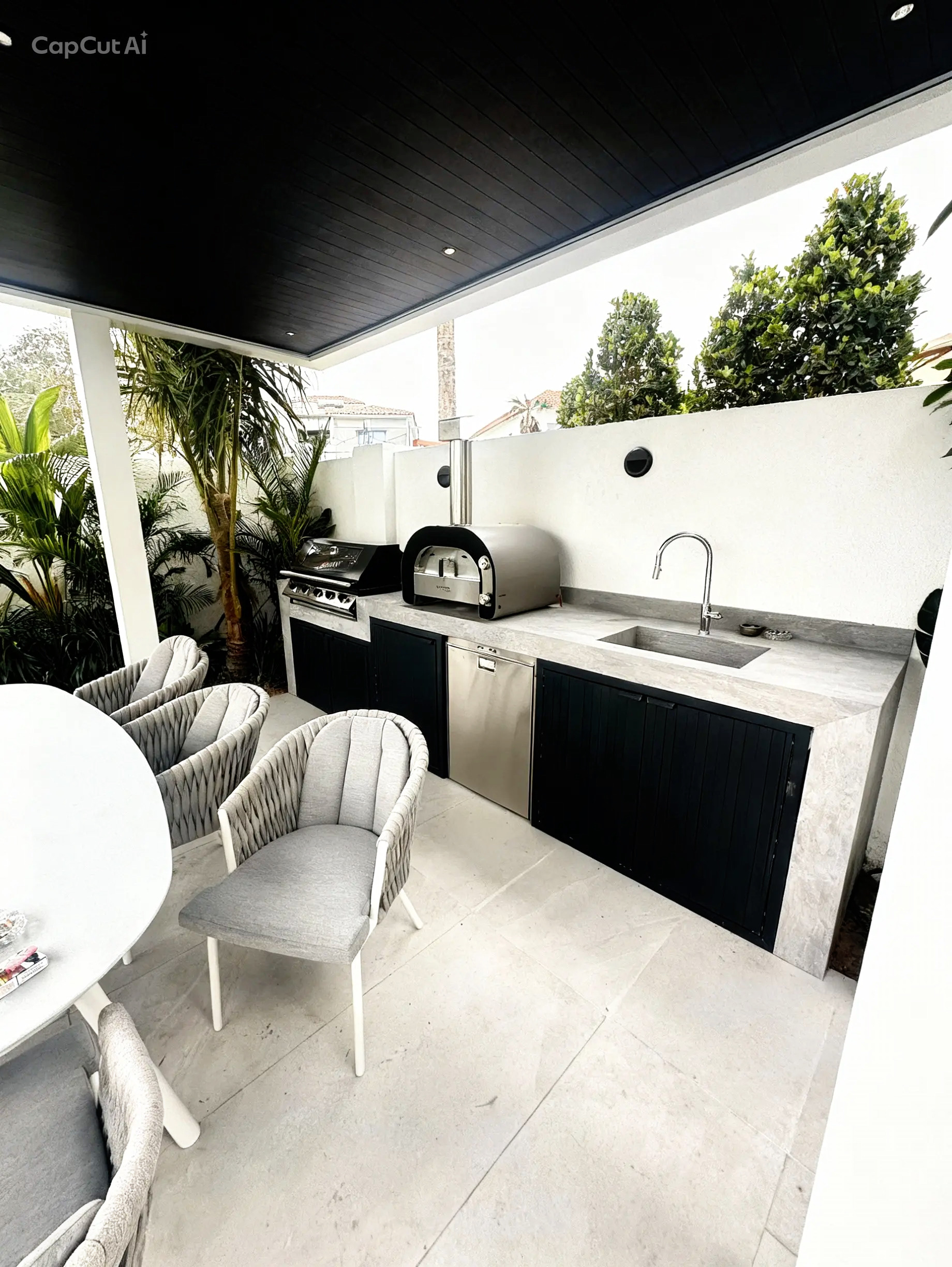 Narrow outdoor seating area with tall green plants along the left side, artificial grass floor, white high tables and stools, and two tall stainless steel patio heaters.